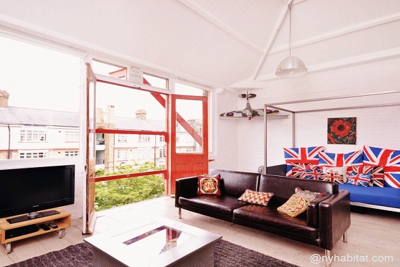 Image of living room of LN-573 with British flag pillows on a couch and large open windows overlooking typical English buildings