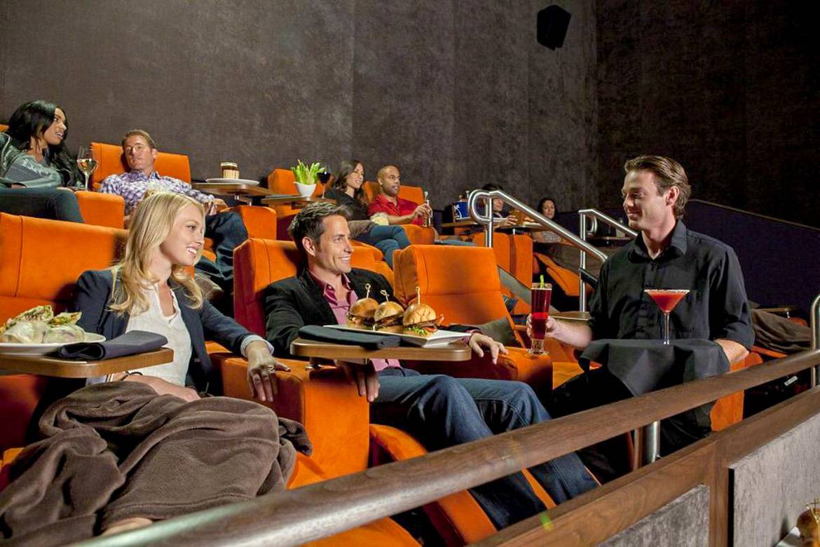 Image of people in a movie theater having dinner and being served cocktails while watching a movie