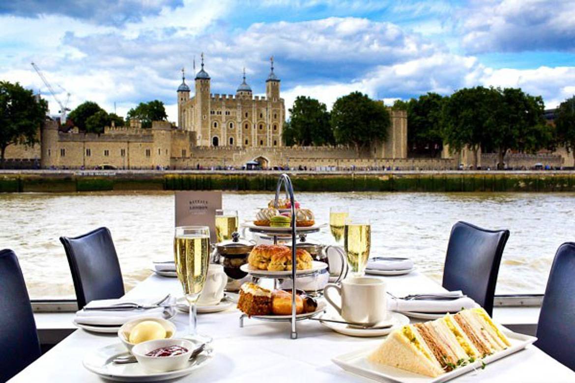 Image of teacups, champagne glasses, sandwiches and scones on a 3-tiered serving tray with a river in the background