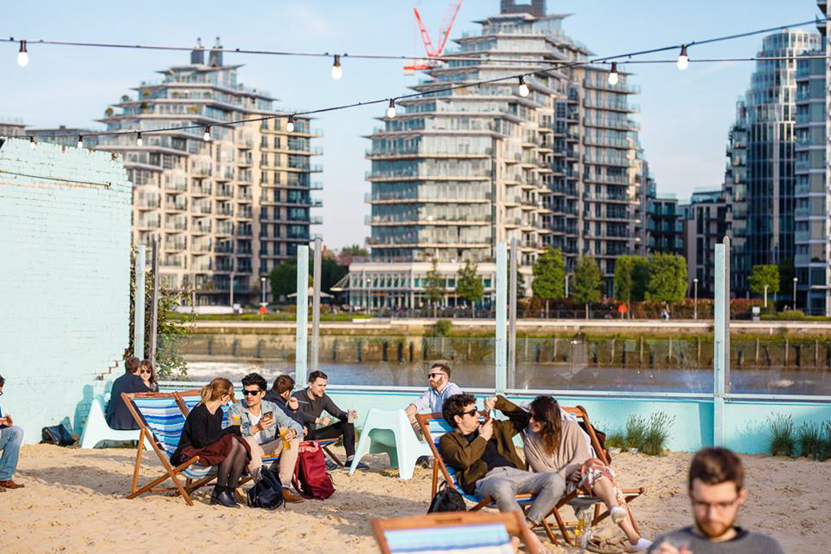 Image of people sitting in lounge chairs at Neverland London’s Fulham beach club with the Thames river in the background.