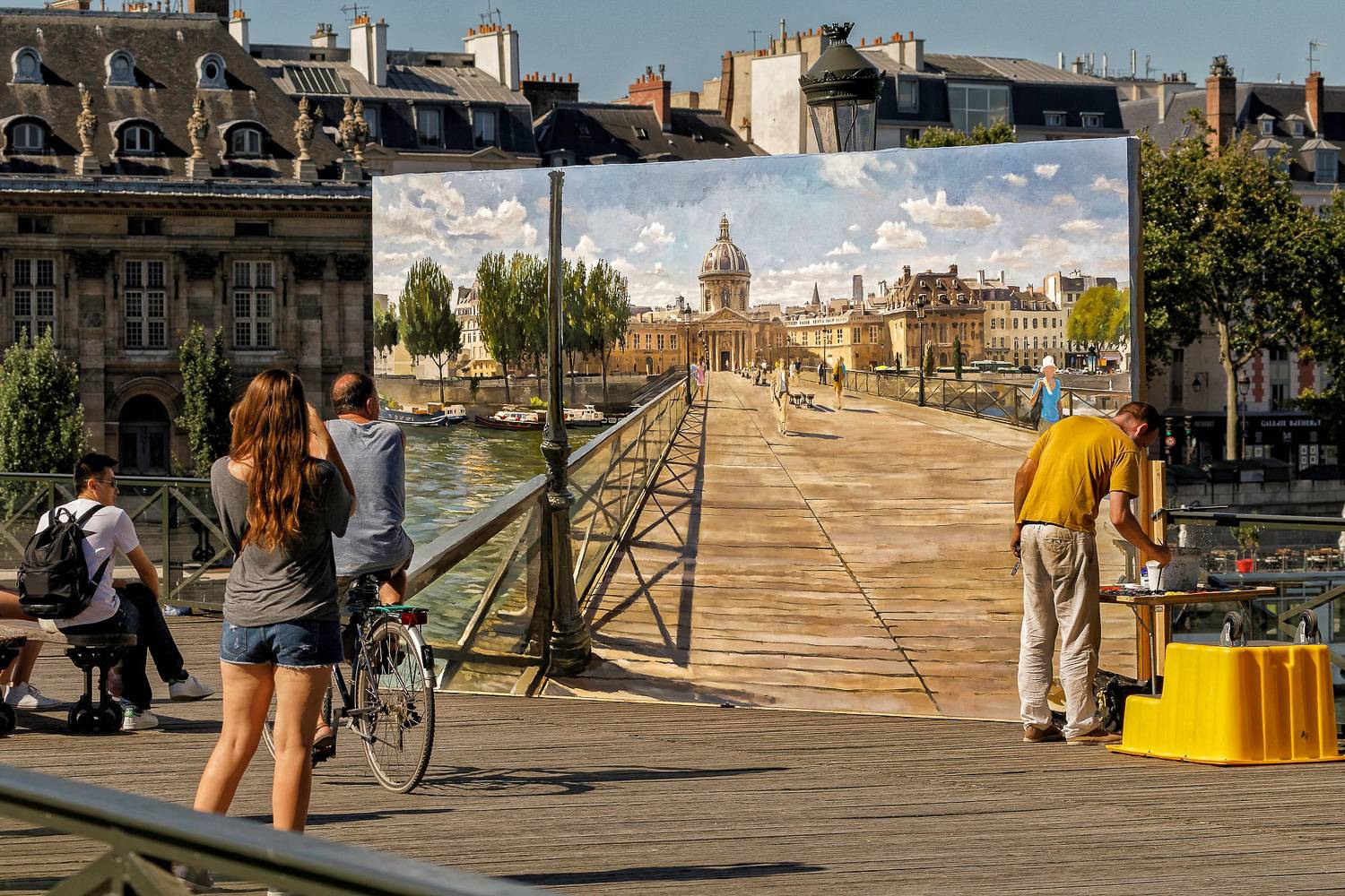 Image of pedestrians and cyclists crossing a bridge over the Seine River while an artist paints the scene.