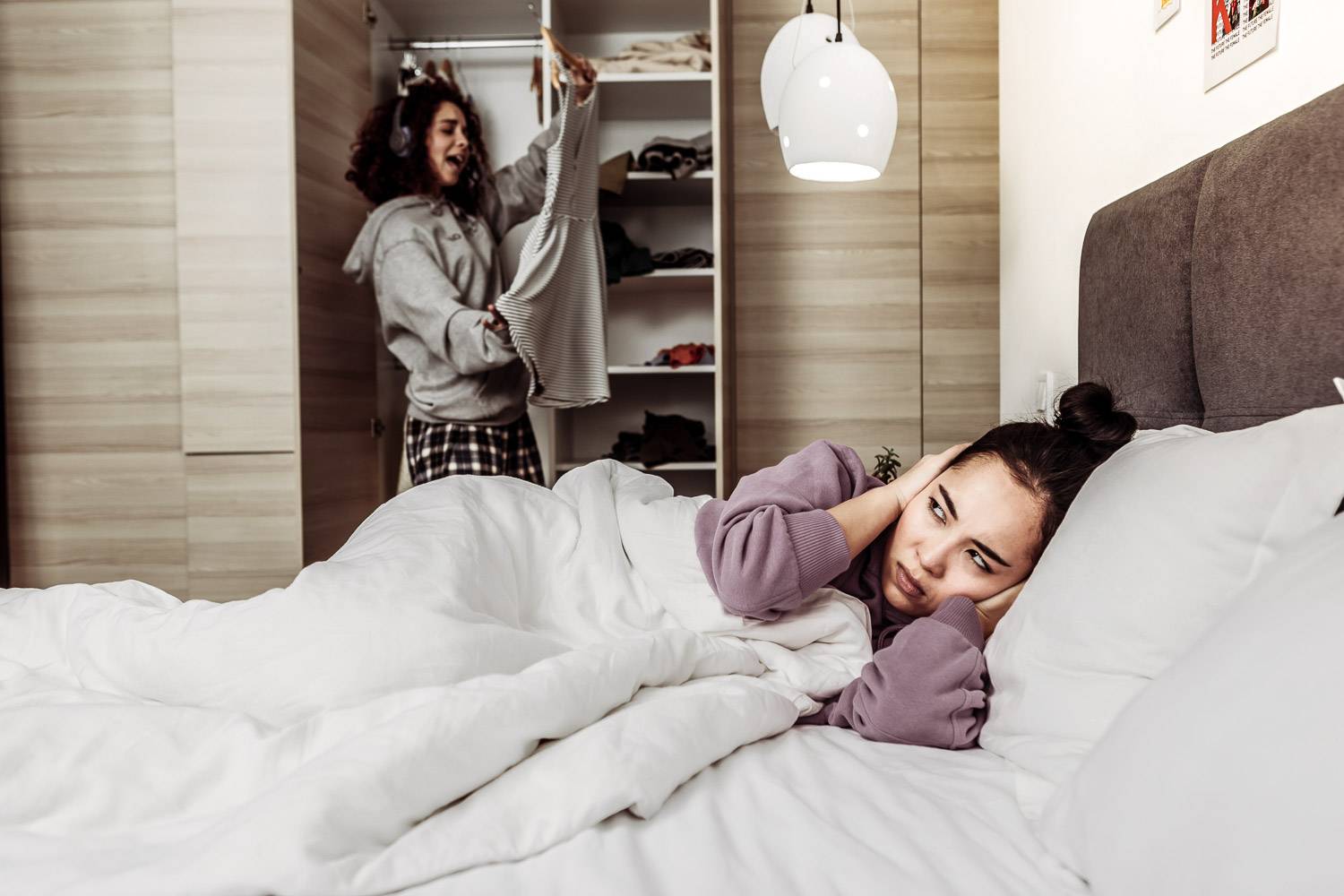 Image of a girl in bed with her hands over her ears while another girl sings with headphones on in the background while looking at clothes in the closet