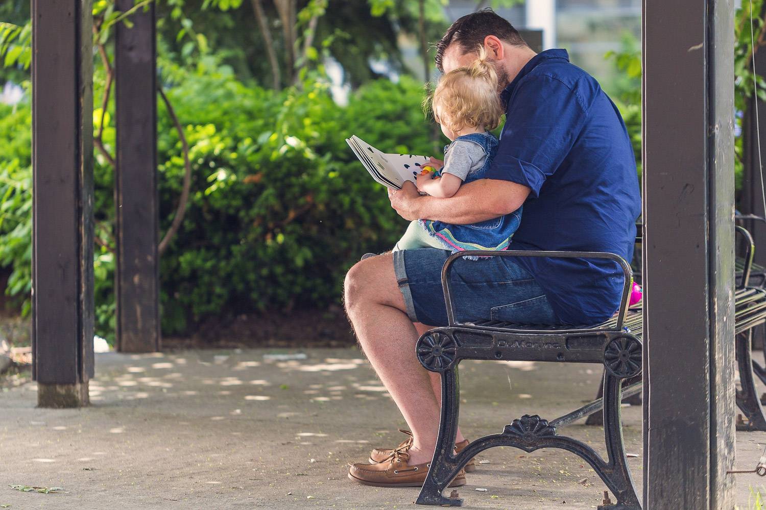 Bild eines Vaters, der mit seinem Baby auf dem Schoss in einem Park sitzt und ein Buch liest
