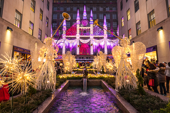 Bild des Rockefeller Centers mit beleuchteten Engeln mit Trompeten, die um einen Brunnen herum stehen, mit Saks Weihnachtsbeleuchtung im Hintergrund