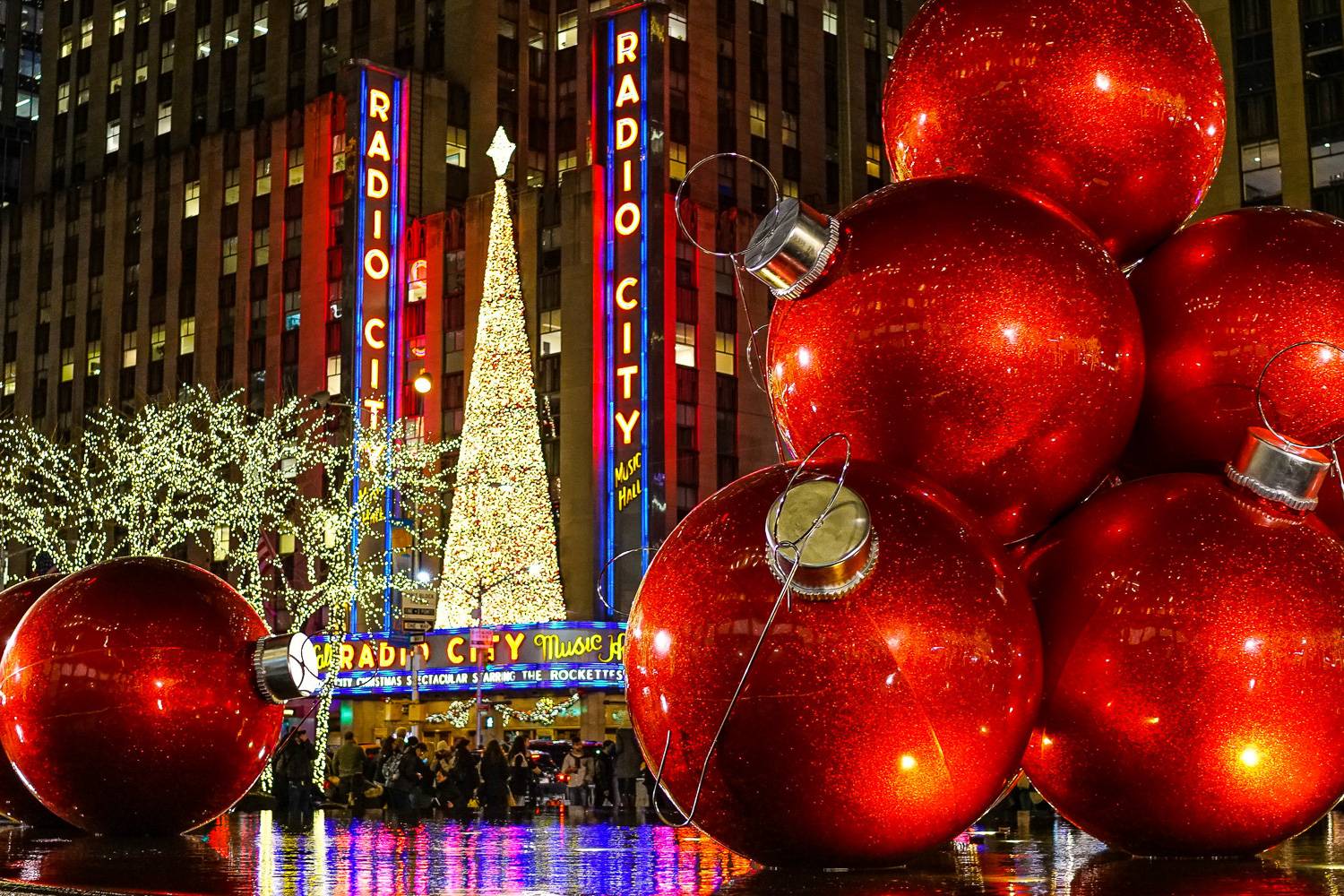 Photo de la devanture du Radio City Music Hall de nuit avec des néons, un sapin de Noël au-dessus de l’entrée, des arbres remplis de guirlandes et des boules de Noël rouges géantes.
