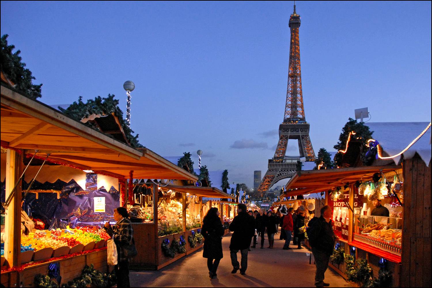 Photo de chalets proposant des produits alimentaires et des cadeaux sur un marché de Noël avec la tour Eiffel en arrière-plan.