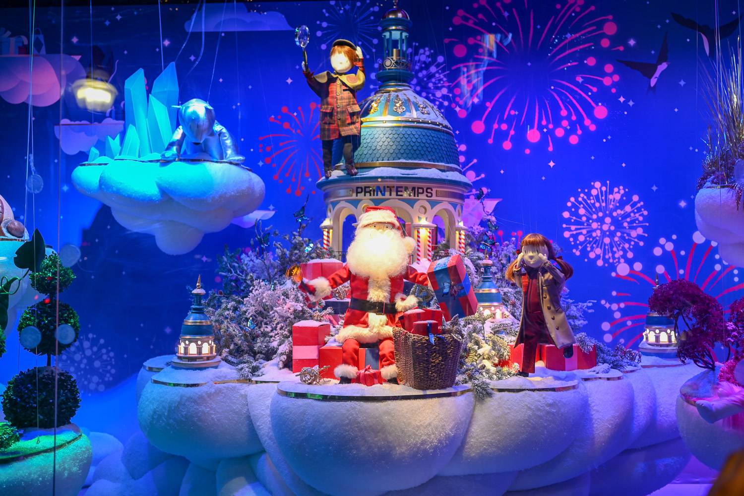 Photo d’une vitrine de Noël entourée de cadeaux, représentant des figurines animées dans un pays merveilleux hivernal, avec le père Noël au milieu.