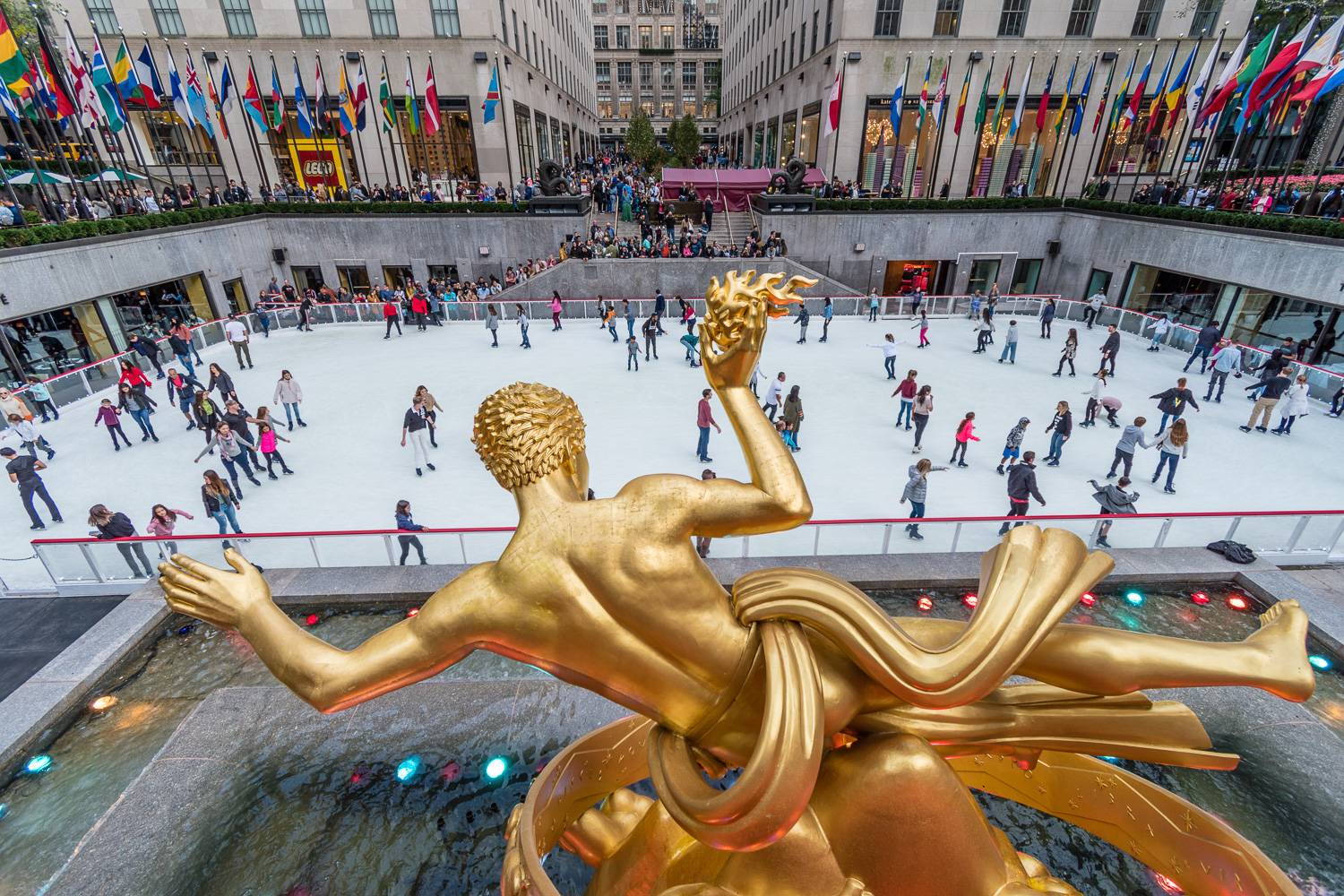 Image de la patinoire du Rockfeller Center avec une statue en or, des drapeaux internatinaux et des personnes en train de patiner (Crédit Photo : Shutterstock)