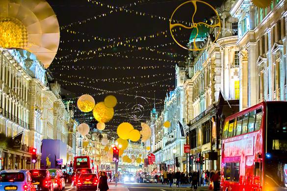 Immagine di una strada con molti negozi con luci festive colorate, autobus a due piani e persone mentre fanno shopping