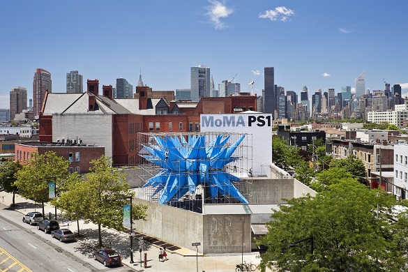 Immagina del MoMa PS1 nel Queens con una scultura blu gigante sul tetto e il panorama di Manhattan sullo sfondo